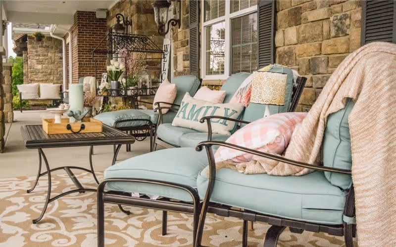 A cozy outdoor patio area with cushioned metal chairs and a loveseat in light blue, decorated with pillows including one that says 'FAMILY'. There is a patterned rug on the floor, a small table with a wooden tray and candles, and a stone wall with windows and shutters in the background.
