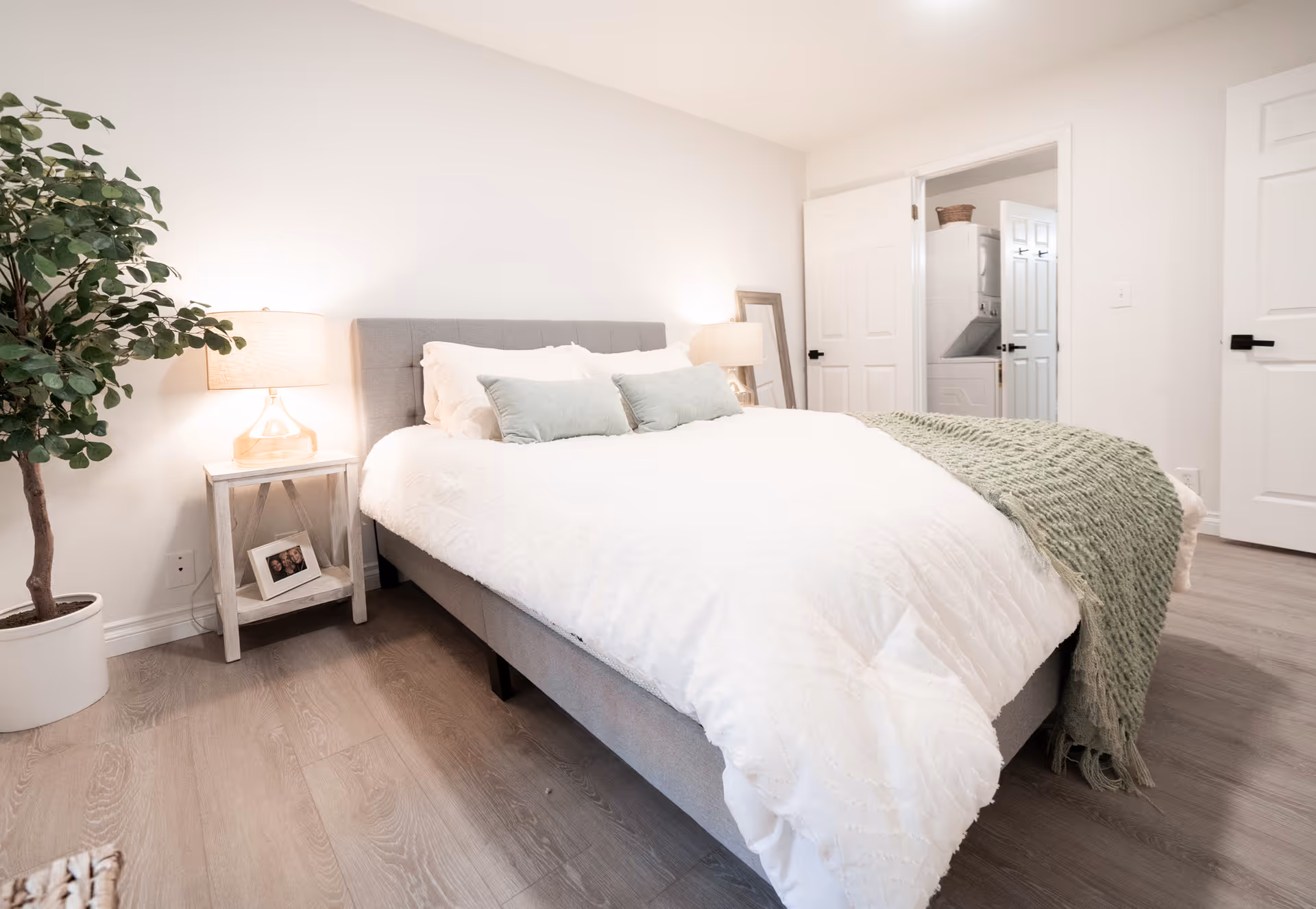 Bright, minimalist bedroom with a bed dressed in white linens, a nightstand and lamp, a potted plant, and an open doorway.