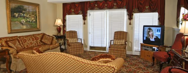 A cozy living room with patterned sofas and armchairs arranged around a carpeted floor. There are three large windows with white blinds and red valances. A television on a wooden stand is turned on, and a table lamp is lit on a side table. A framed painting hangs on the wall above one sofa.
