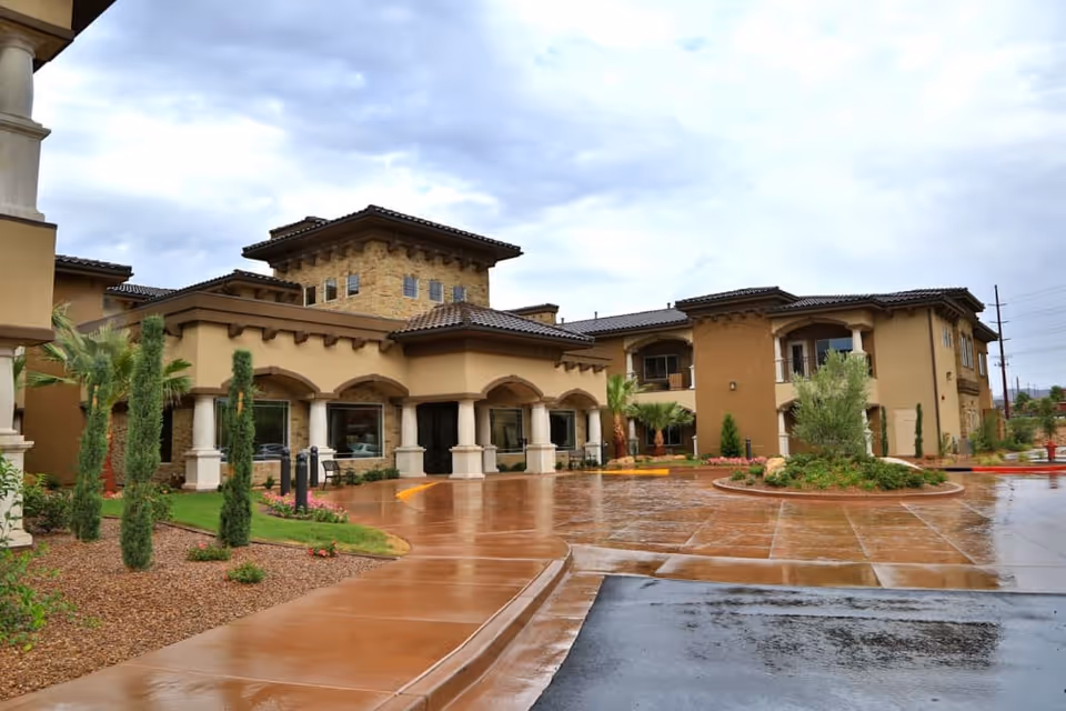Exterior view of The Retreat at Sunbrook senior living facility showing a large building with beige and brown tones, arched windows and doorways, columns, and a tiled roof. The foreground features a wet driveway and landscaped areas with small trees and shrubs under a cloudy sky.