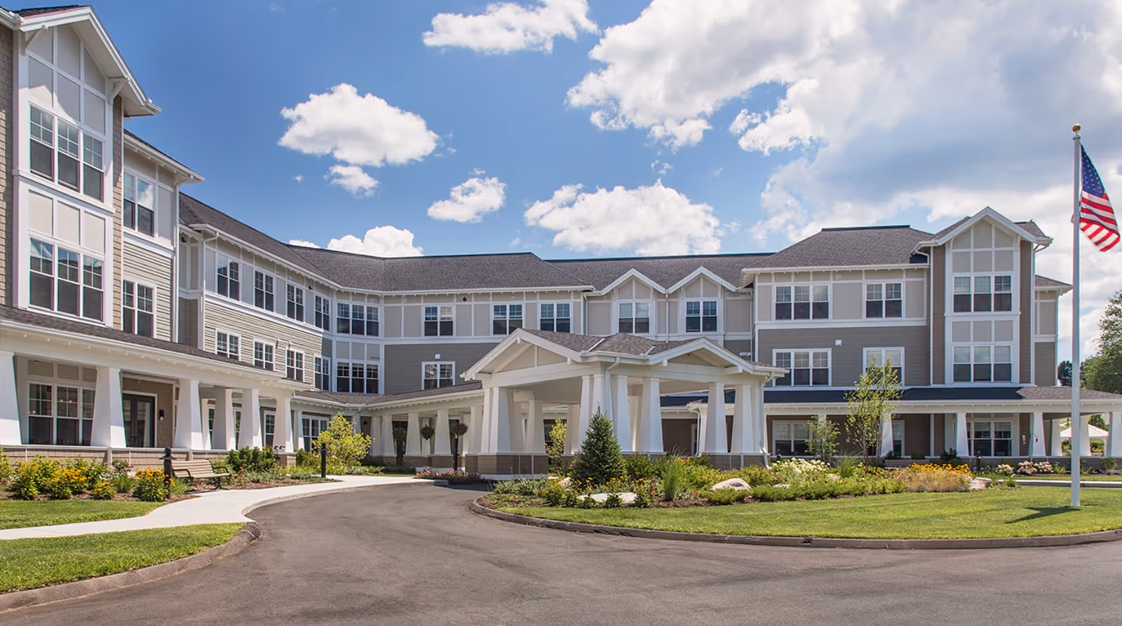 Exterior view of a large, modern senior living facility building with multiple windows, a covered entrance, landscaped gardens, and an American flag on a flagpole under a partly cloudy blue sky.