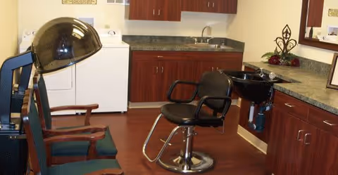 Interior view of a salon area in a senior living facility featuring a black salon chair in front of a black hair washing sink, two green cushioned waiting chairs, a large black salon dryer, and cabinetry with a countertop and sink in the background.