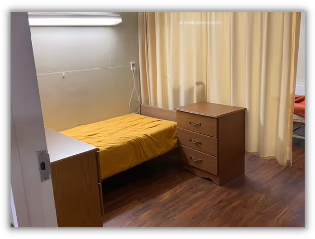 A small room with a single bed covered in a yellow blanket, a wooden three-drawer dresser next to the bed, and a curtain divider separating this space from another area with a bed visible behind the curtain. The floor is wooden, and the walls are light-colored.