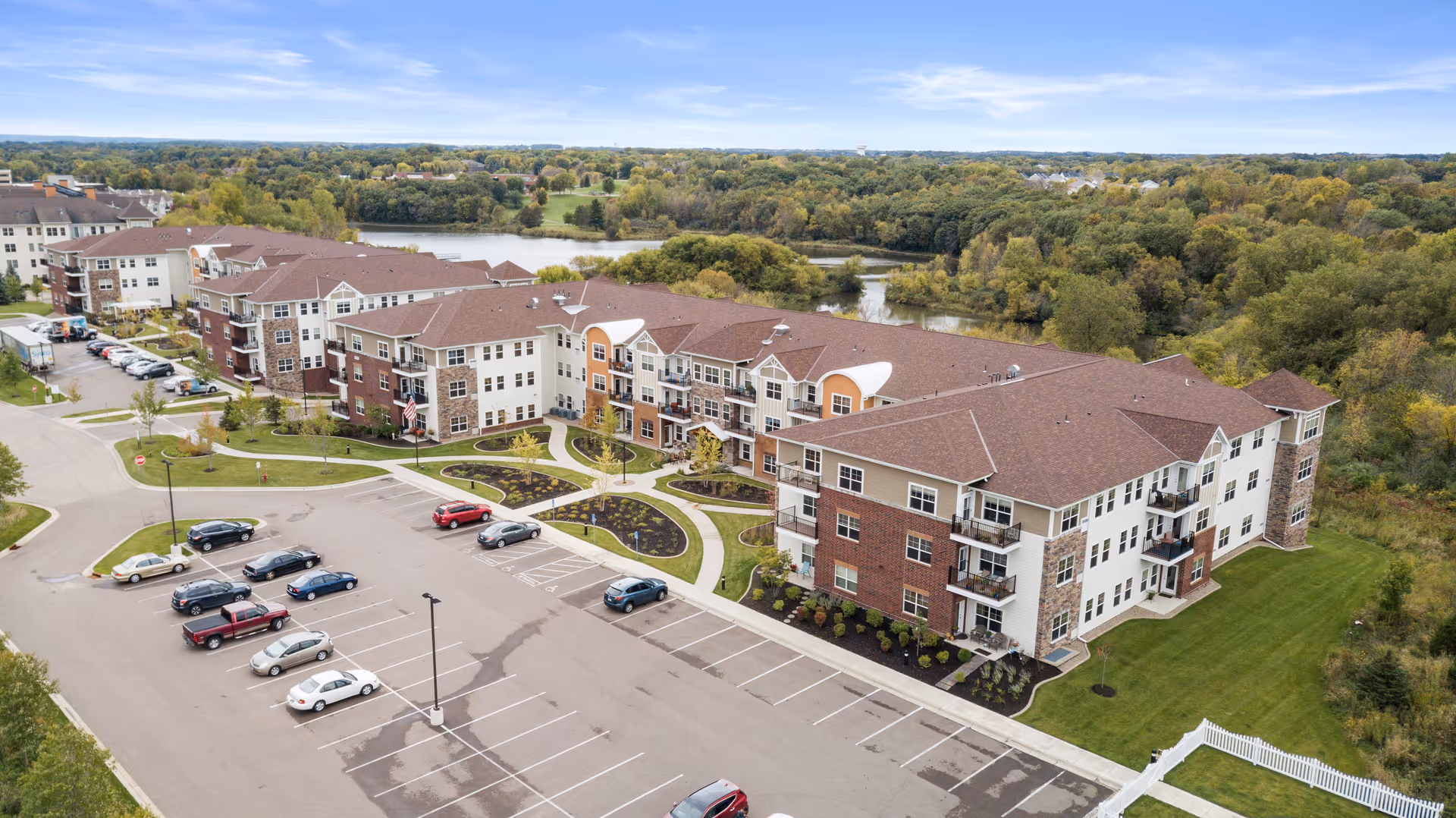 Aerial view of the Chaska Heights senior living complex showing multi-story buildings, landscaped courtyards, a parking lot, and surrounding woods with a pond.