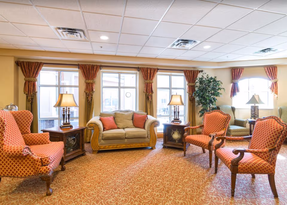 Sunlit sitting room with a sofa, patterned armchairs, side tables and lamps arranged on a patterned carpet.