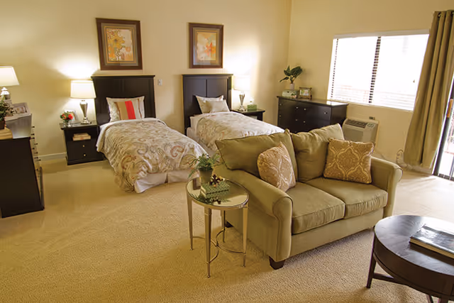 A well-lit bedroom with two twin beds featuring patterned bedspreads and decorative pillows. Between the beds is a nightstand with a lamp and small decor items. In front of the beds is a green loveseat with two patterned cushions and a small round glass-top side table with a plant. The room also includes a dark dresser under a window with curtains and an air conditioning unit. The walls are light-colored, and there are framed pictures above the beds.