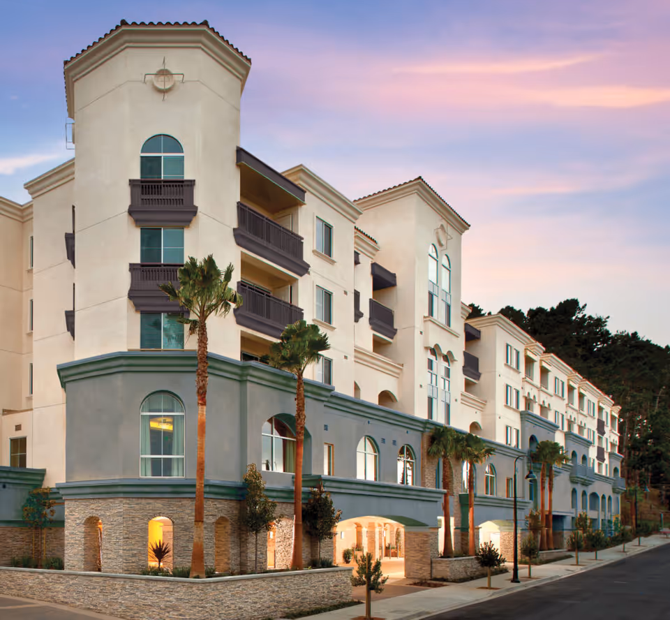 Exterior view of the multi-story Peninsula Del Rey senior living building with balconies, arched windows, palm trees, and a pastel evening sky.