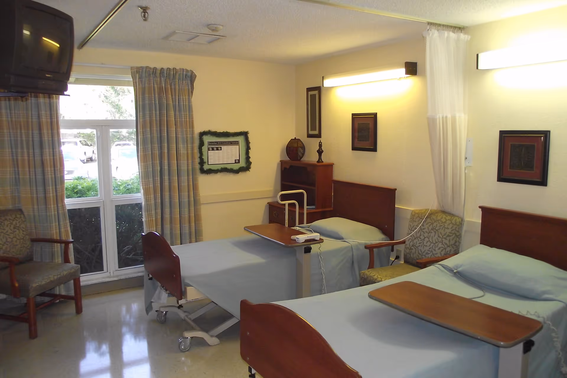 A room in a rehabilitation and nursing center with two hospital beds, each equipped with an overbed table. The beds have light blue sheets and are positioned against the walls. There are two upholstered chairs, a window with plaid curtains, framed pictures on the walls, and a small wooden cabinet between the beds. The room is softly lit with wall-mounted lights.