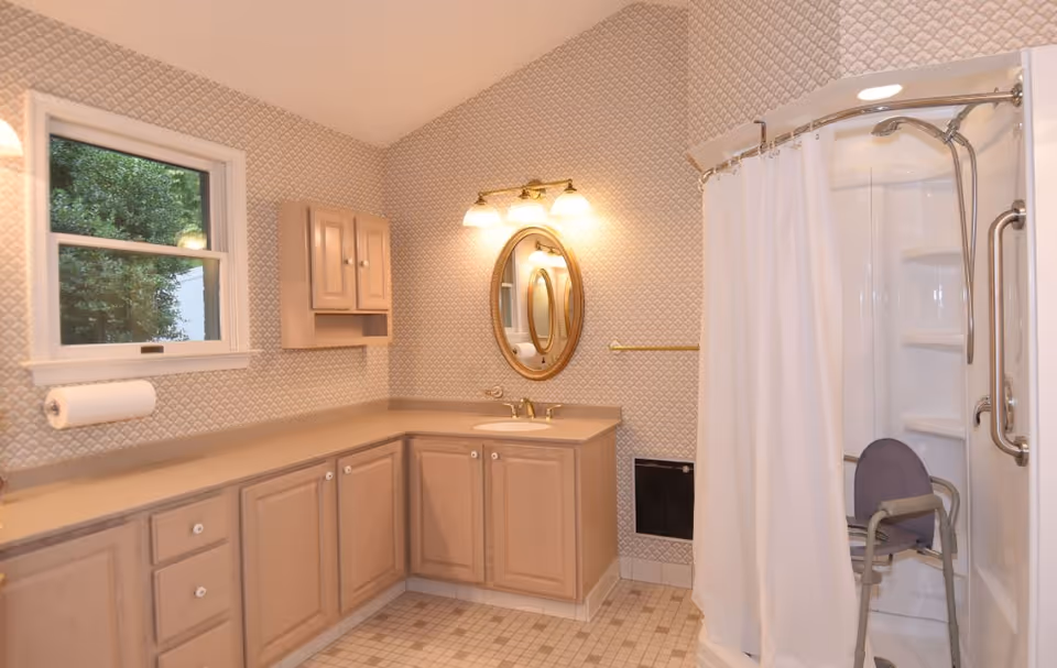A bathroom with beige cabinetry and a beige countertop. There is a round mirror above the sink with three light fixtures above it. A window is on the left wall, and a paper towel holder is mounted below it. On the right side, there is a shower with a white curtain and a shower chair inside. The walls have a patterned wallpaper, and the floor is tiled with small square tiles.