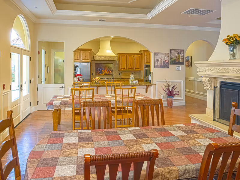 A cozy dining area with wooden tables covered in patchwork-patterned tablecloths and matching wooden chairs. In the background, there is a kitchen with wooden cabinets, a stove with a decorative hood, and a refrigerator. The room features a fireplace with an ornate mantel on the right side and a vase with flowers. The walls are painted light beige, and there is a large arched window and door letting in natural light.