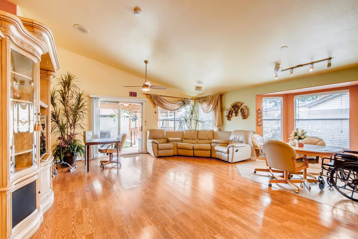 A bright and spacious living room with wooden flooring, a beige leather sectional sofa, a round table with chairs, a wheelchair, large windows with blinds, and sliding glass doors leading to an outdoor area. The room also features a wooden cabinet, potted plants, ceiling fan, and decorative wall hangings.
