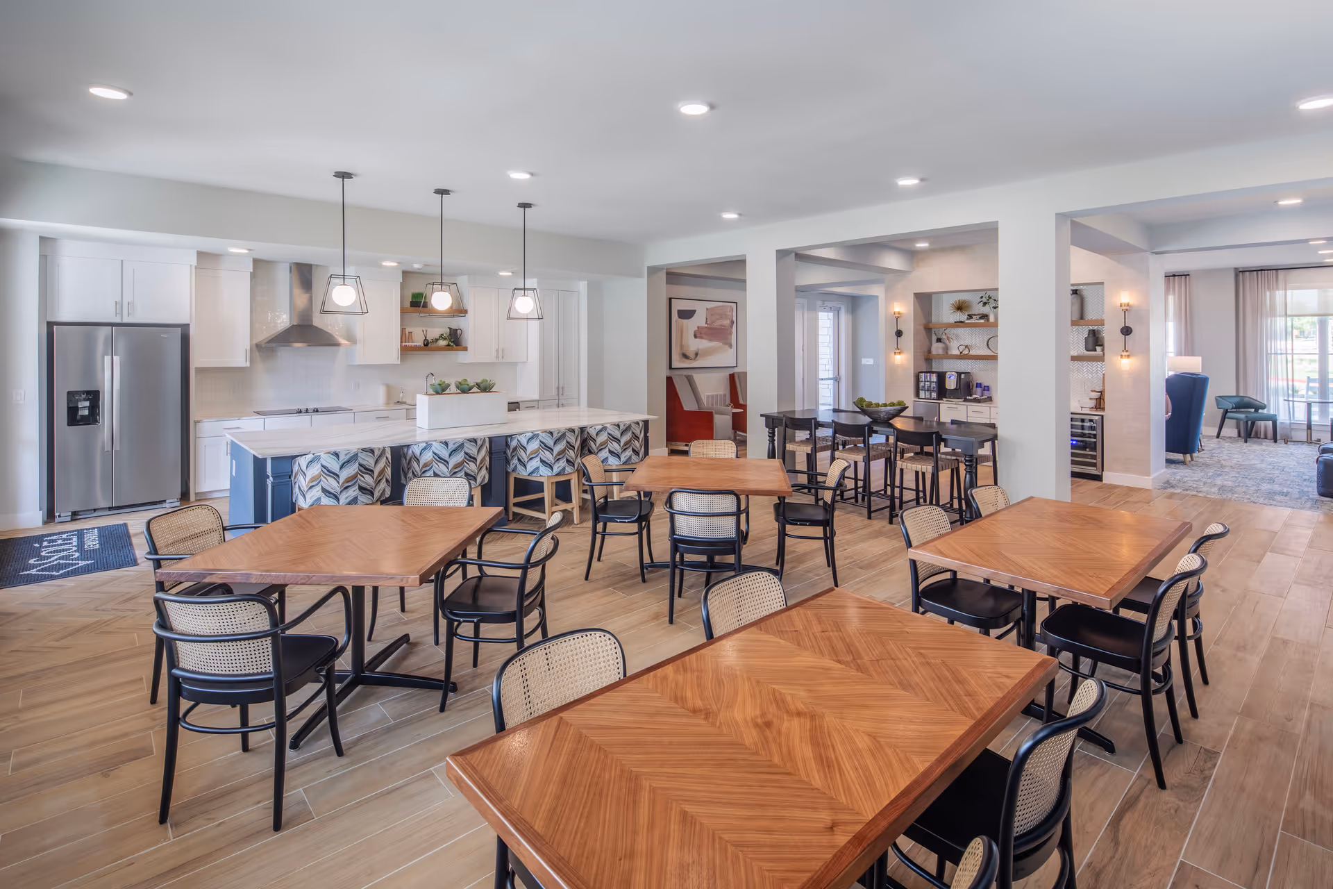 Open communal dining and kitchen area with multiple wooden tables and chairs, a large island with pendant lights, and lounge seating in the background.