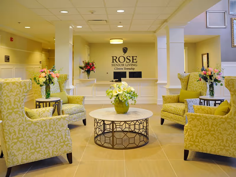 A bright and welcoming senior living facility lobby with four patterned yellow armchairs arranged around a round coffee table with a green vase of white flowers. Behind the seating area is a reception desk with two computer monitors and a wall sign that reads 'ROSE SENIOR LIVING Clinton Township'. There are additional flower arrangements on side tables and the reception desk.