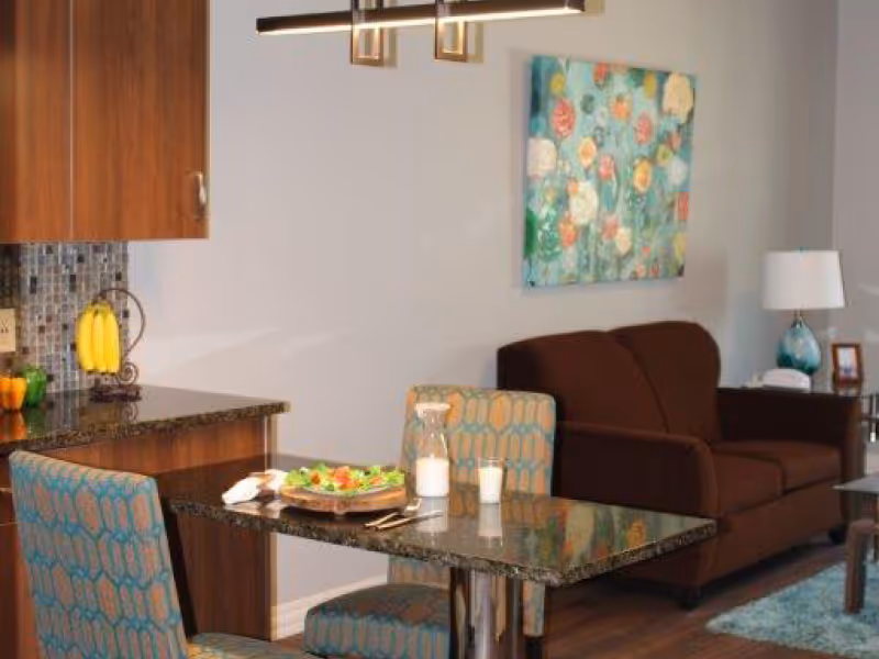 A cozy interior space featuring a small dining area with a granite table set for a meal, two patterned chairs, a brown sofa, a side table with a lamp, and a colorful floral painting on the wall.