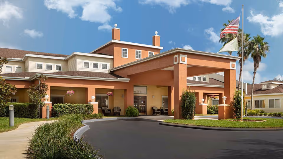 Front exterior view of Colliers Assisted Living at Port St. Lucie showing a large covered entrance with seating areas, well-maintained landscaping, and two flagpoles with the American flag and another flag against a blue sky with some clouds.