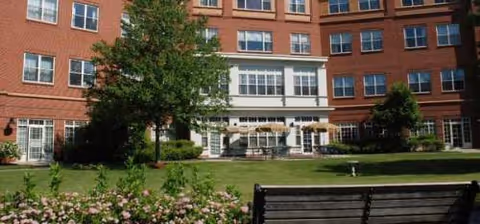 A brick senior living building exterior with a white entrance and bench on a landscaped lawn.