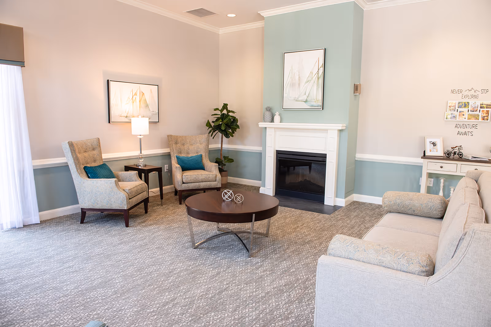 A cozy living room with a light gray sofa, two patterned armchairs with teal cushions, a round wooden coffee table with decorative spheres, a white fireplace with a painting above it, a small side table with a lamp, a potted plant, and a console table with framed photos and wall art that reads 'Never Stop Exploring Adventure Awaits'.