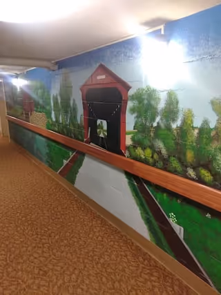 Indoor hallway with a carpeted floor and a handrail on the right side. The wall features a colorful mural depicting a covered bridge, trees, and a river scene. The hallway is lit by ceiling lights.