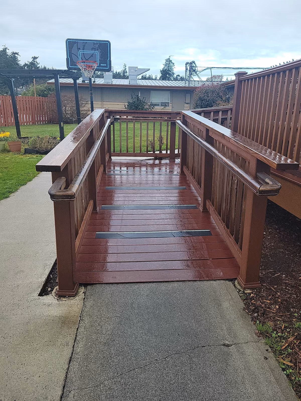 A wooden wheelchair ramp with handrails on both sides leading down to a concrete pathway. In the background, there is a basketball hoop, a grassy area, and a building with windows and a metal roof. The ramp and surrounding area appear wet from recent rain.
