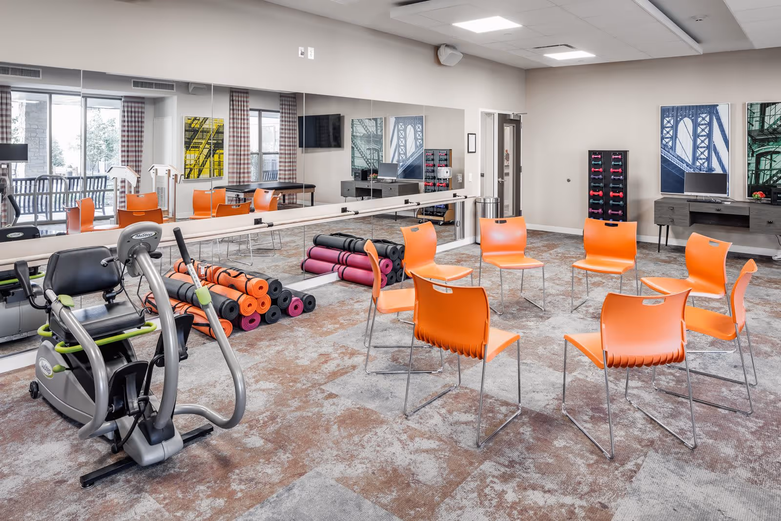 A fitness and activity room with eight orange chairs arranged in a circle, exercise equipment including a stationary bike, and several rolled-up yoga mats stacked against a mirrored wall. The room has large windows with checkered curtains, colorful artwork on the walls, and a desk with a computer and dumbbell rack in the background.