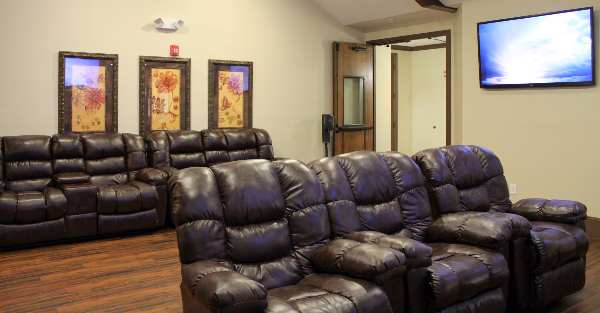 A seating area with rows of dark leather recliner sofas facing a wall-mounted TV and floral artwork on the wall.