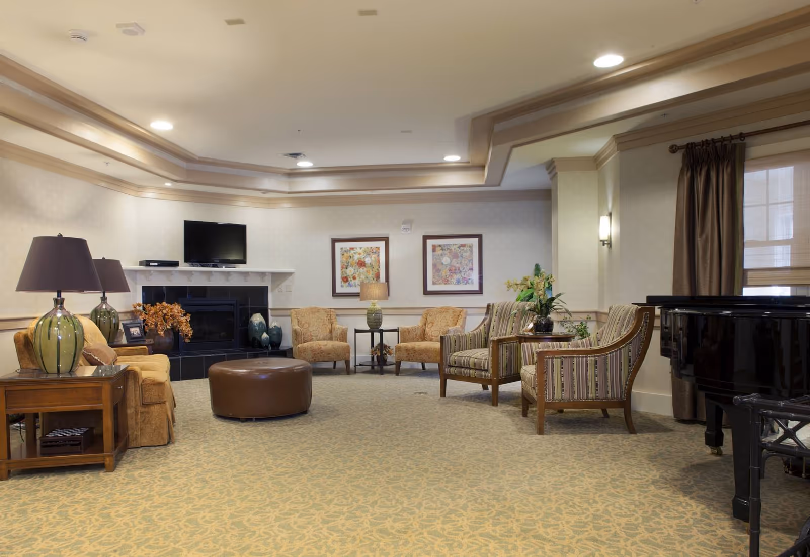 A spacious living room in a senior living facility with beige walls and carpet. The room features a fireplace with a TV mounted above it, two framed floral artworks on the wall, multiple upholstered chairs and sofas arranged around a round brown ottoman, side tables with lamps, and a black grand piano near a window with brown curtains.