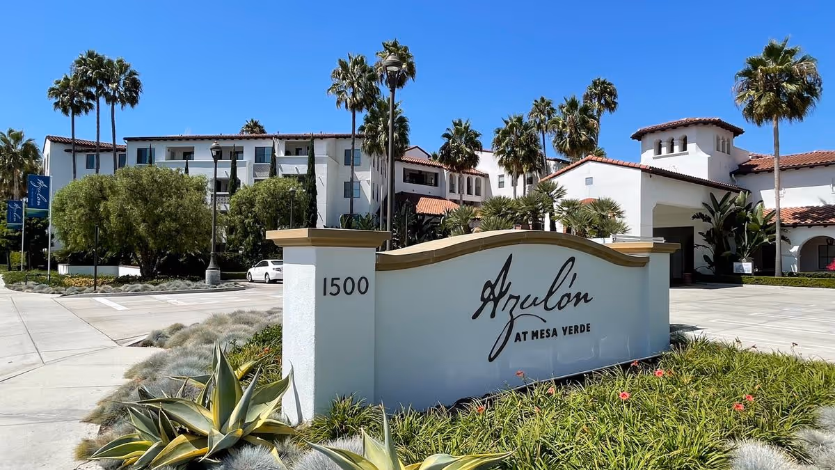 Entrance sign for Azulon at Mesa Verde in landscaped grounds with a Mediterranean-style building and palm trees behind it.