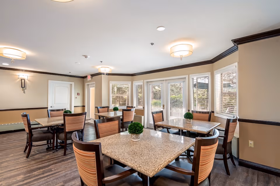 Bright communal dining room with multiple square tables and wooden chairs, large windows and glass doors letting in natural light.