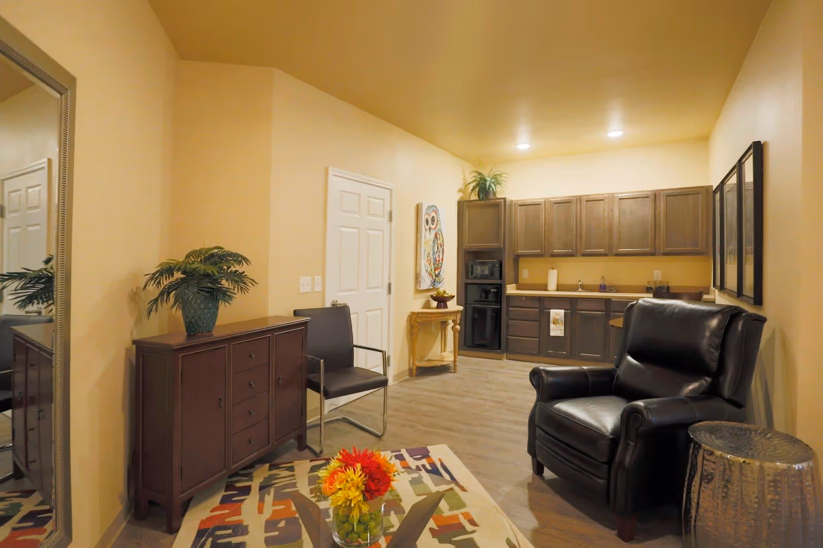 A cozy interior room with beige walls and wooden flooring featuring a small kitchenette with upper and lower cabinets, a sink, and a microwave. There is a black leather armchair, a black chair with metal arms, a wooden cabinet with a potted plant, a small wooden table with a decorative owl painting above it, and a colorful patterned rug with a glass vase of flowers on a glass coffee table.