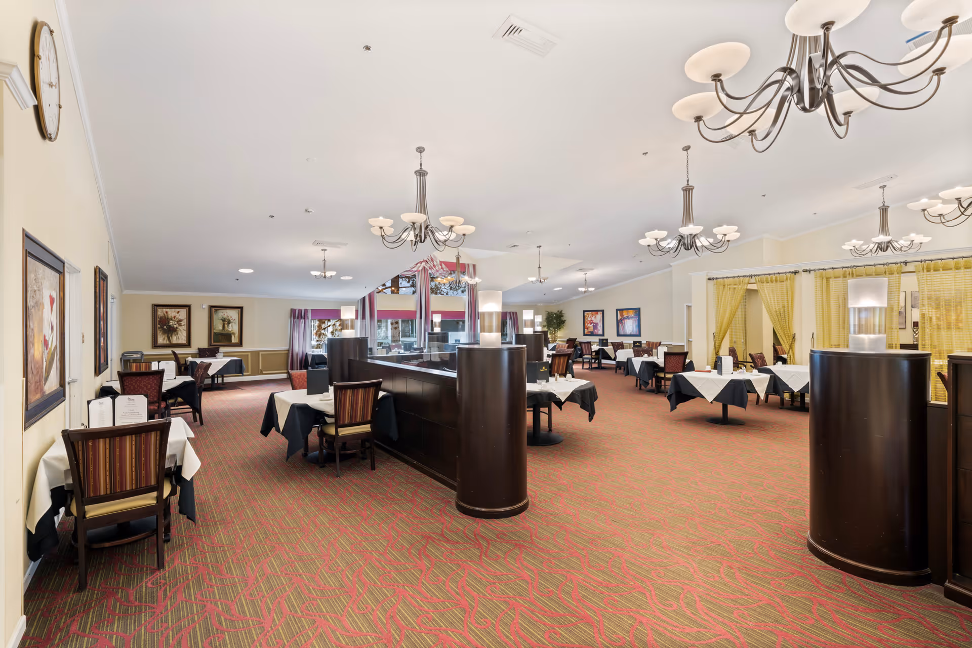 Spacious dining room with multiple tables covered in white and black tablecloths, surrounded by wooden chairs with striped cushions. The room features chandeliers hanging from the ceiling, framed artwork on the walls, and large windows with curtains allowing natural light to enter.