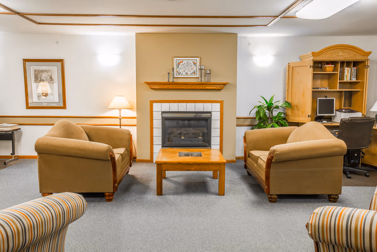 A cozy living room area with two beige upholstered sofas facing each other across a wooden coffee table. In the center background, there is a fireplace with a wooden mantle decorated with small items and a framed picture above it. To the right, there is a wooden cabinet with an open door revealing a computer and office chair. A green potted plant is next to the cabinet. The room has soft lighting from wall sconces and a floor lamp, with a framed picture on the left wall.