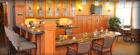 Interior view of a senior living facility dining area with a long granite countertop bar surrounded by chairs. The background features wooden cabinetry with glass doors, a wall-mounted TV, and large windows letting in natural light. There are small tables and chairs arranged near the windows and a potted plant in the corner.