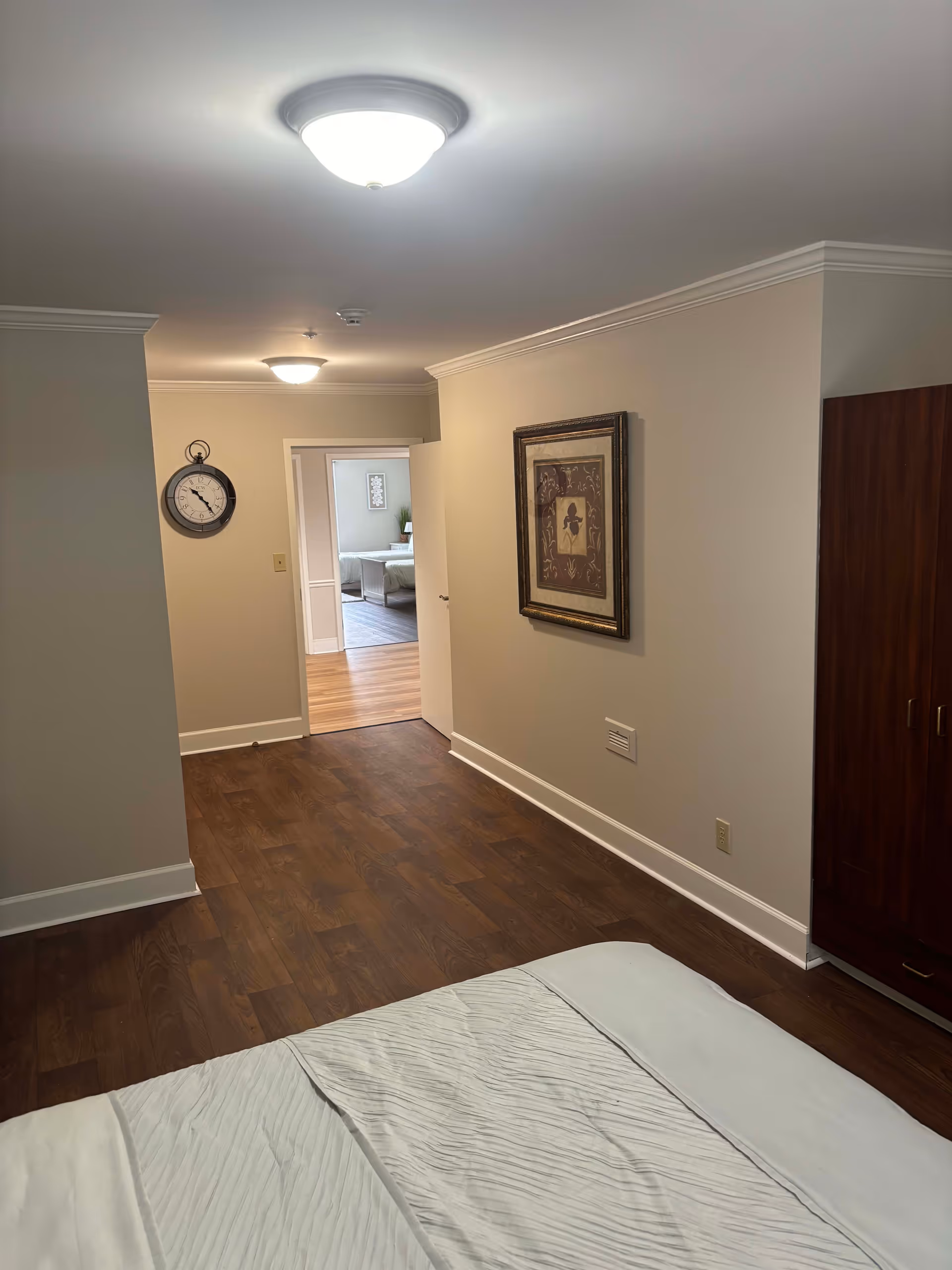 View of a senior living facility bedroom with a bed partially visible in the foreground, wooden flooring, beige walls, a framed picture on the wall, a wooden wardrobe, and a hallway leading to another room with a clock on the wall.
