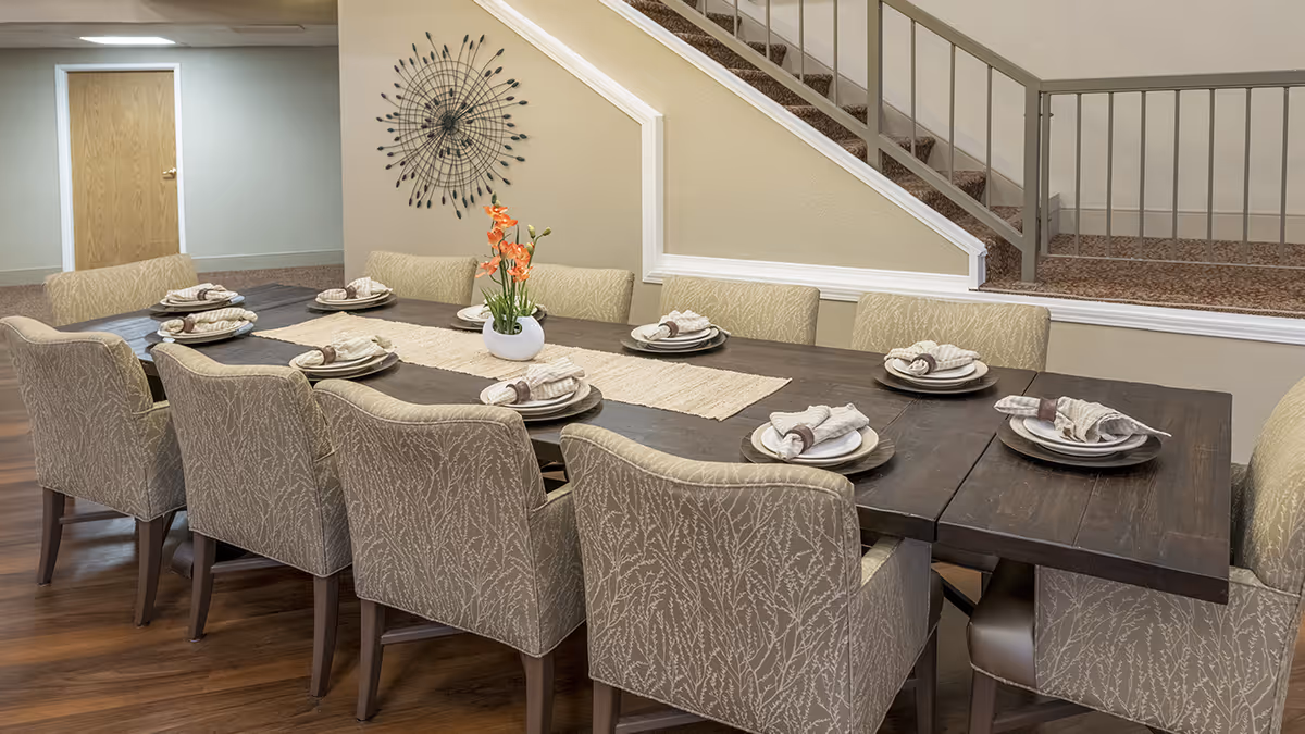 A long wooden dining table set with plates, napkins, and napkin rings, surrounded by upholstered chairs with a leafy pattern. A small white vase with orange flowers is placed in the center of the table. The room has wooden flooring, beige walls, a decorative wall clock, and a staircase with a metal railing in the background.