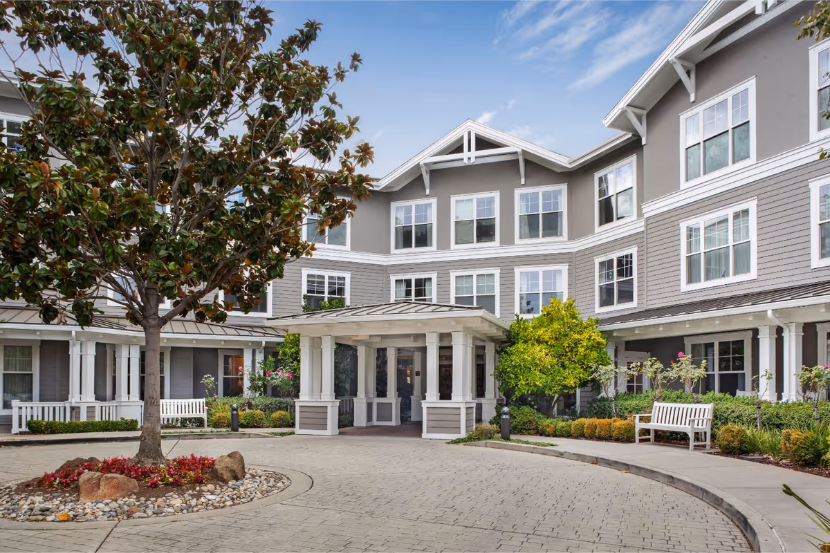 Exterior view of Ivy Park at Belmont senior living facility showing a three-story building with gray and white siding, multiple windows, a covered entrance with white pillars, landscaped garden with trees, bushes, flowers, and benches along a paved driveway.