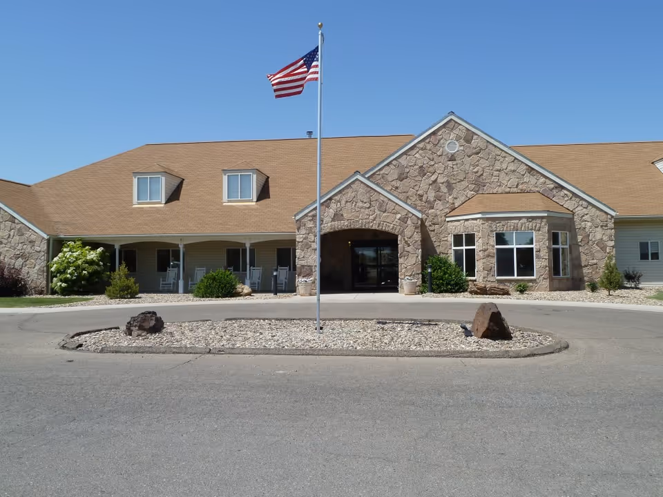 Front exterior view of Evergreen Gardens facility with a stone facade, a tan roof, and an American flag on a flagpole in the center of a circular driveway. There are bushes and a few rocks in the landscaped area around the flagpole.