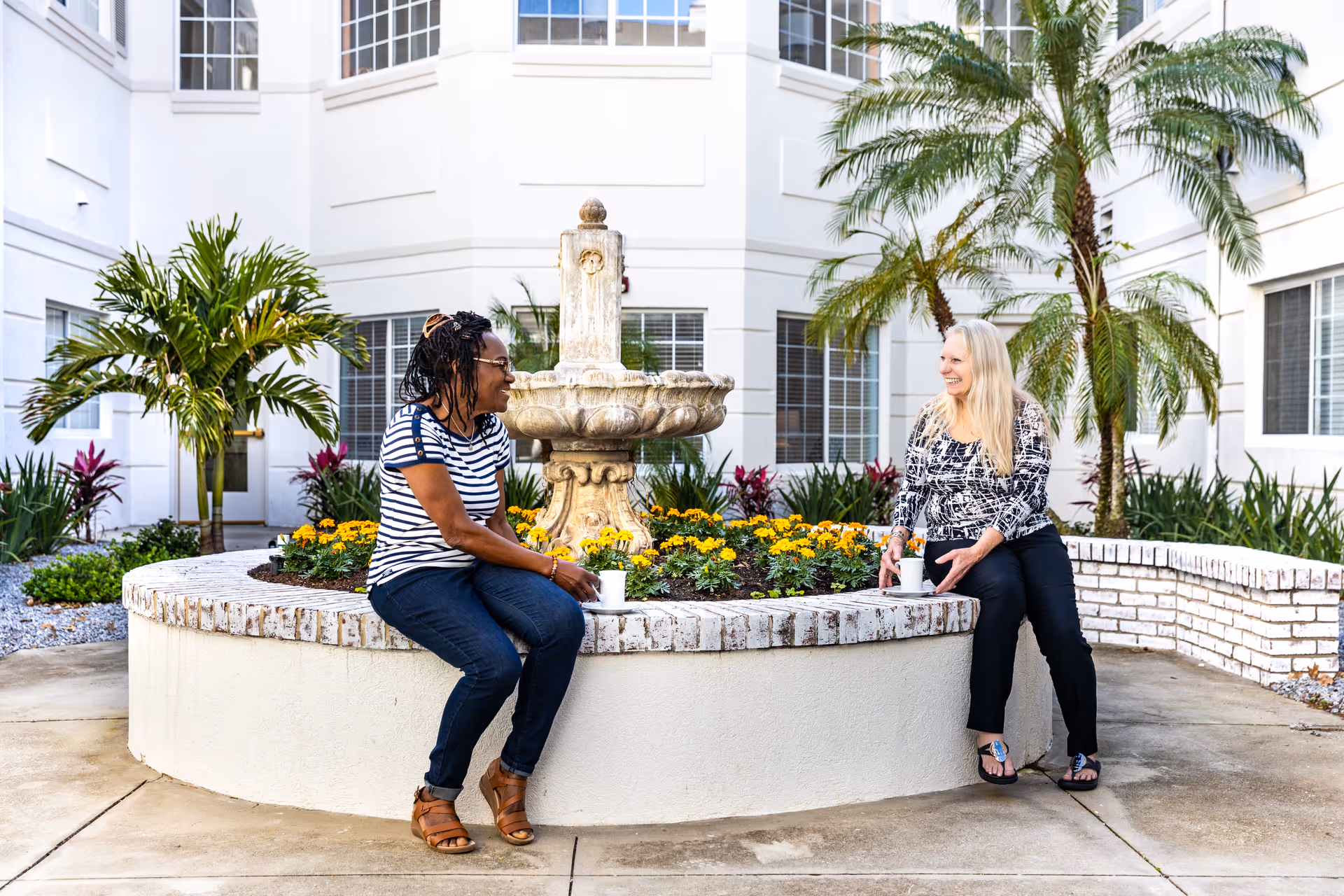 Two women sit and chat on the edge of a circular planter with a fountain, flowers, and palm trees in a sunny courtyard.
