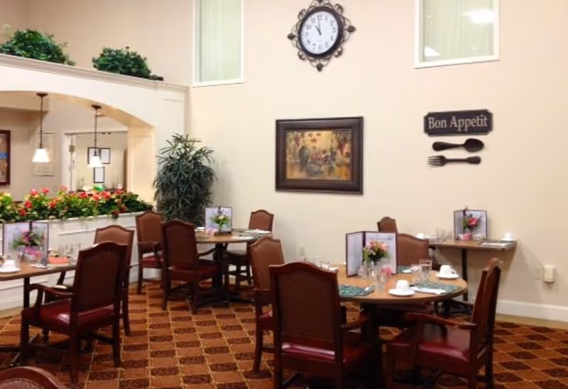 Dining area with several round tables set with menus, cups, and glasses. The room has brown cushioned chairs, a patterned carpet, a wall clock, a framed painting, and a decorative sign that says 'Bon Appetit' with a fork and spoon on the wall. There are plants and flowers near a half wall with an archway leading to another room.