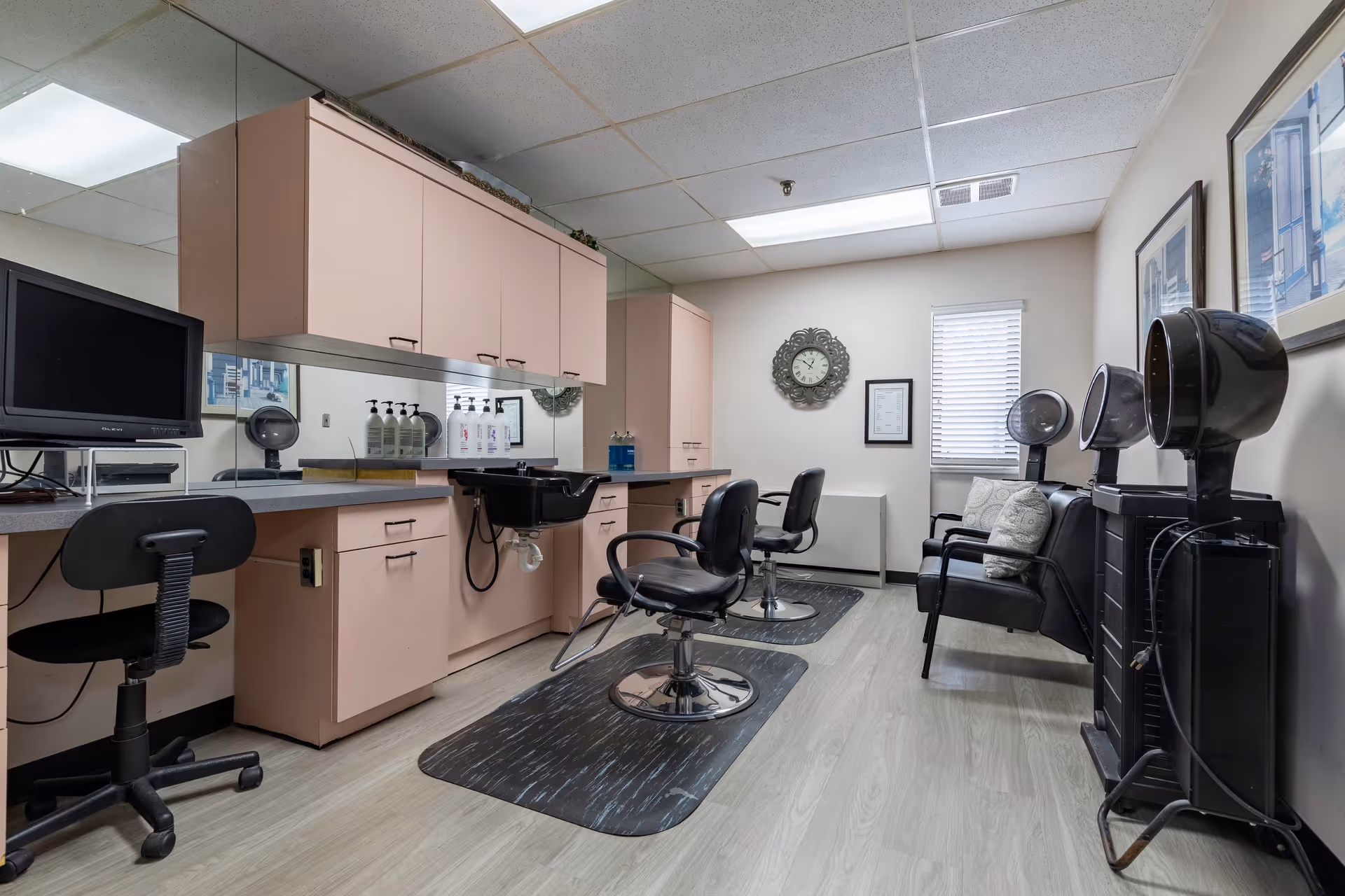 Interior view of a hair salon area in a senior living facility with two black salon chairs in front of a counter with a sink, cabinets, and a mirror. There are hair dryers on the right side and a small seating area with two black chairs and pillows. A clock and framed pictures hang on the walls.
