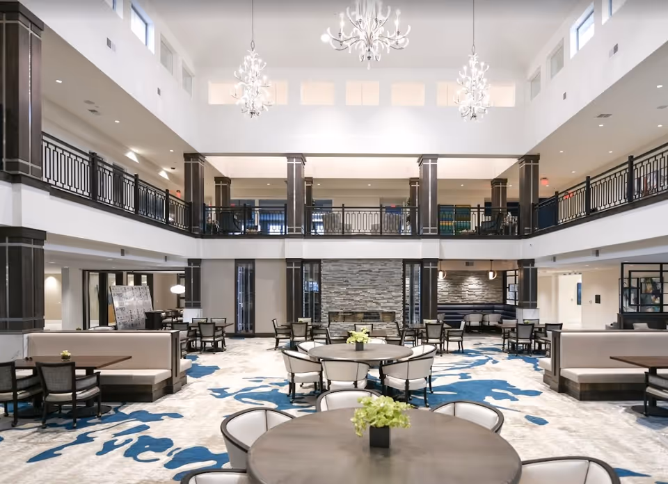 Spacious and modern common area with round tables and chairs arranged on a carpet with blue and beige patterns. The room features a high ceiling with three chandeliers, a second-floor balcony with black railings, and a stone fireplace on the far wall. The space is well-lit with natural and artificial light.