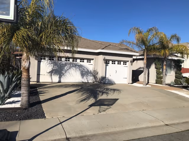 Single-story residential building with three white garage doors, palm trees, and a concrete driveway under a clear blue sky.
