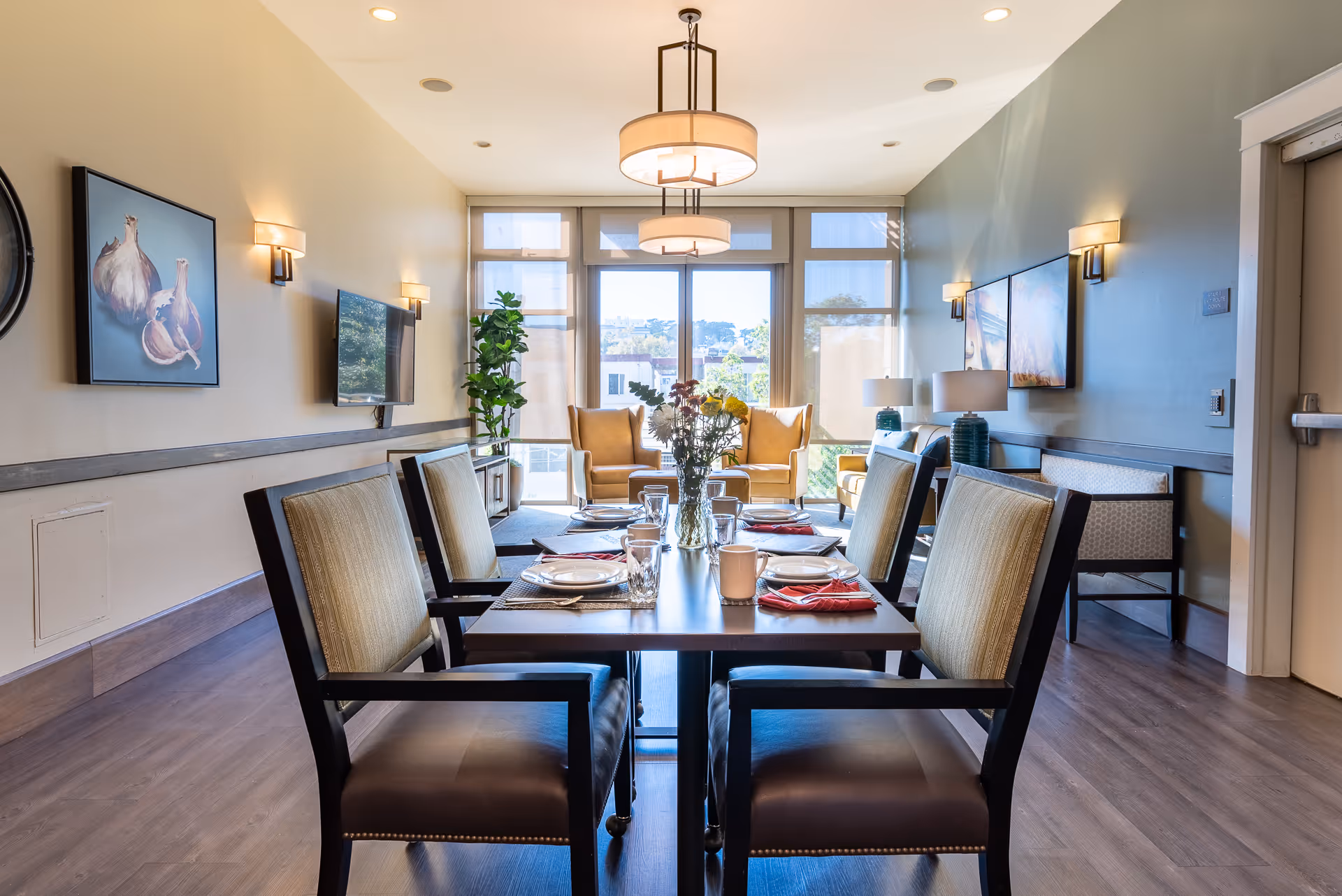 A bright dining room with a rectangular table set for six people, featuring plates, glasses, and red napkins. The room has wooden flooring, beige walls, and large windows letting in natural light. There are two yellow armchairs near the windows, a TV mounted on the wall, and decorative paintings and lamps on the walls.