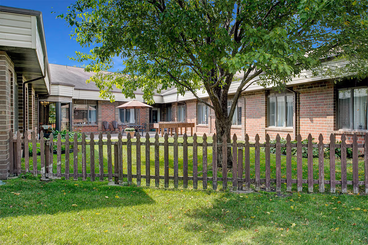 A fenced outdoor courtyard area with a large tree in the center, surrounded by a brick building with multiple windows. There is a patio with outdoor furniture including chairs and a table with an umbrella, under a clear blue sky.