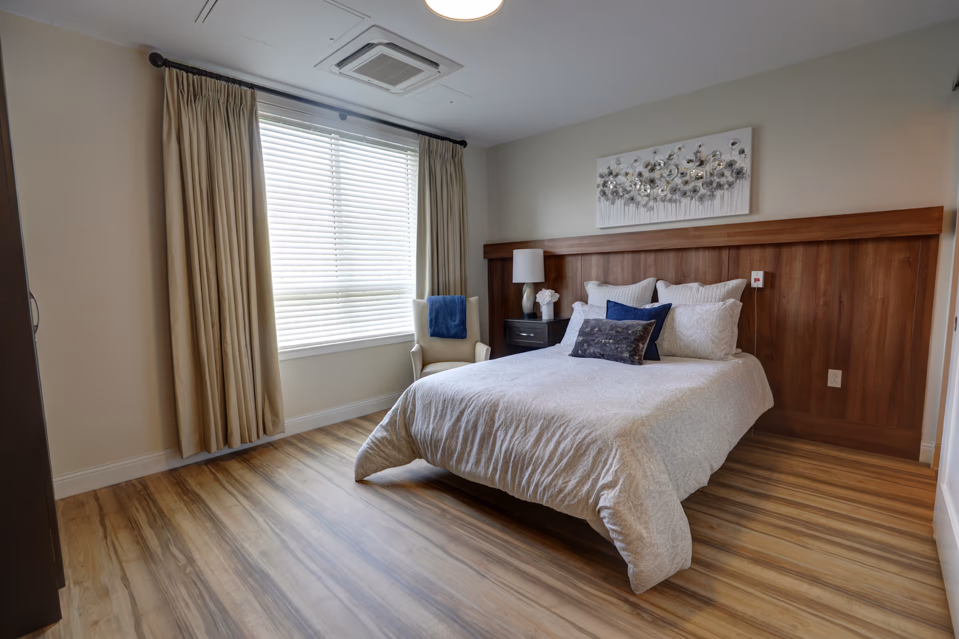 Bright bedroom with a made double bed against a wooden headboard, a nightstand and a chair by a large window with curtains.