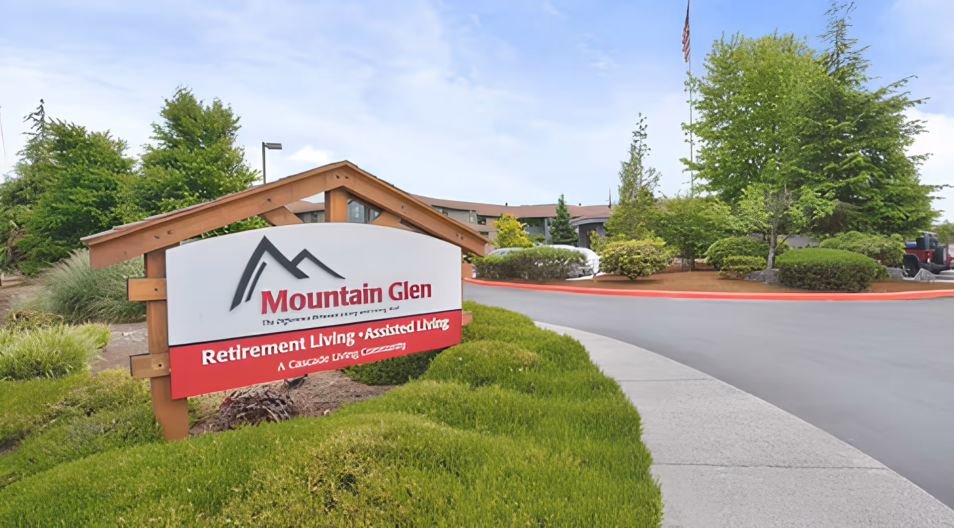 Outdoor view of the entrance to Mountain Glen Retirement and Assisted Living facility, featuring a wooden sign with the facility's name and surrounded by green bushes and trees under a partly cloudy sky.
