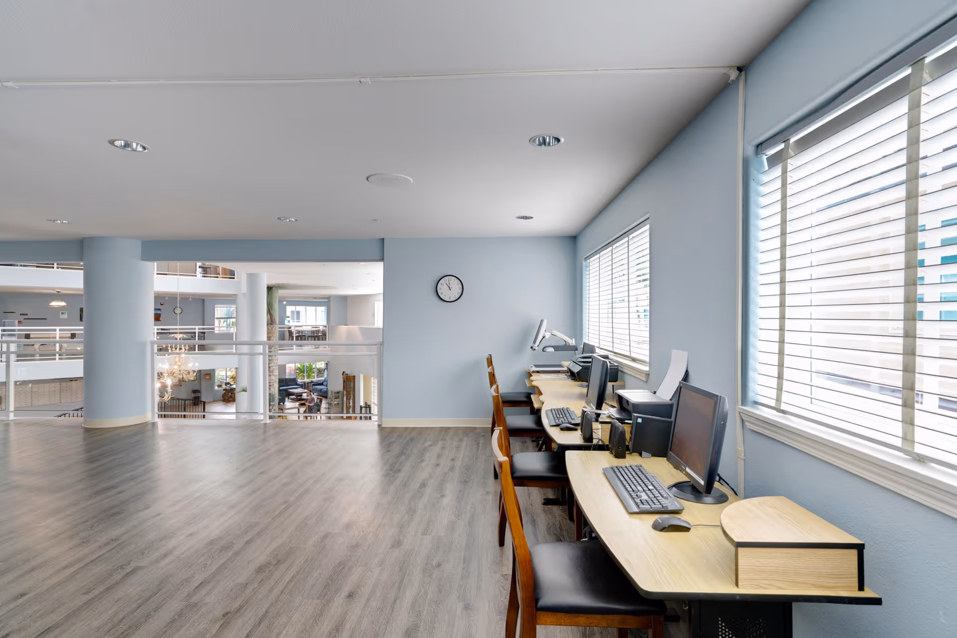 A bright, spacious interior area with light blue walls and wood flooring. On the right side, there is a row of desks with computers, keyboards, and chairs positioned in front of large windows with white blinds. A clock is mounted on the wall above the desks. The left side opens up to a view of a lower floor with railings and additional seating areas visible.