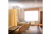 A hospital or nursing center room with two beds separated by a yellow privacy curtain. The room has a large window with blinds, a wooden chair, and a small table. The walls are light-colored with a wooden trim.