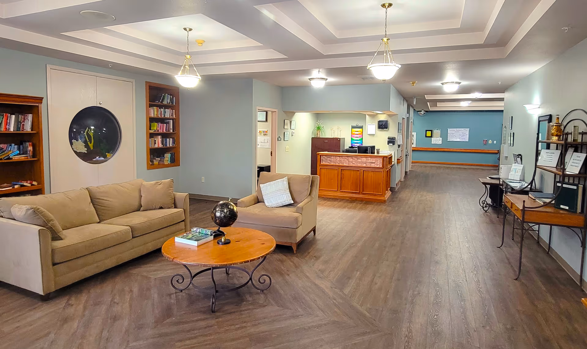 A spacious senior living facility lobby with beige sofas, a round wooden coffee table, built-in bookshelves, and a front desk along a hallway.