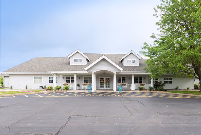 Single-story senior living building with a covered central entrance, front porch seating, a parking lot, and a tree in front.