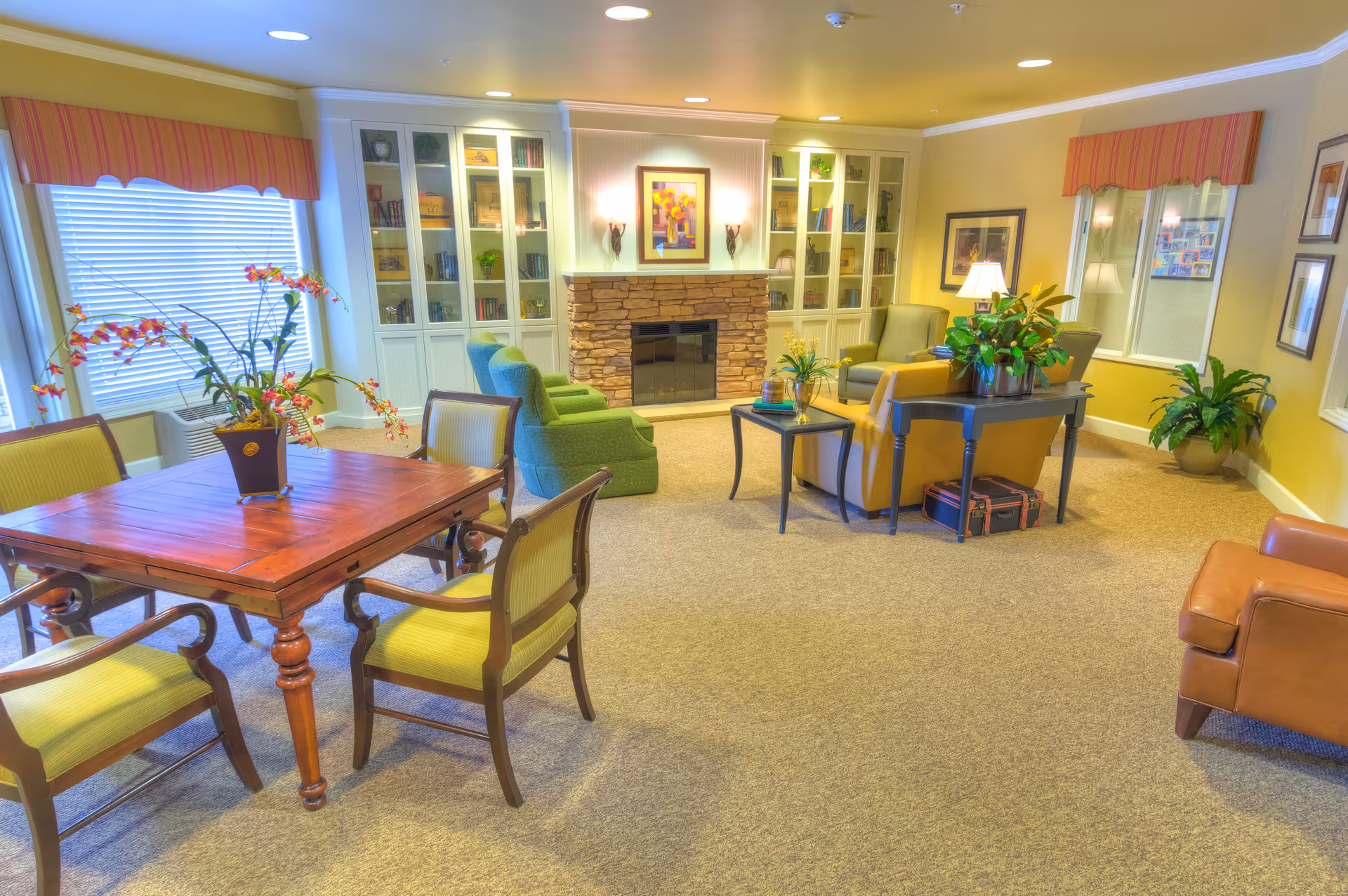 Bright communal living room with armchairs, a wooden table and chairs, built-in bookcases, and a stone fireplace.